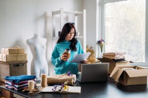 A woman is seated at a table, working on her eCommerce site on a laptop surrounded by boxes.